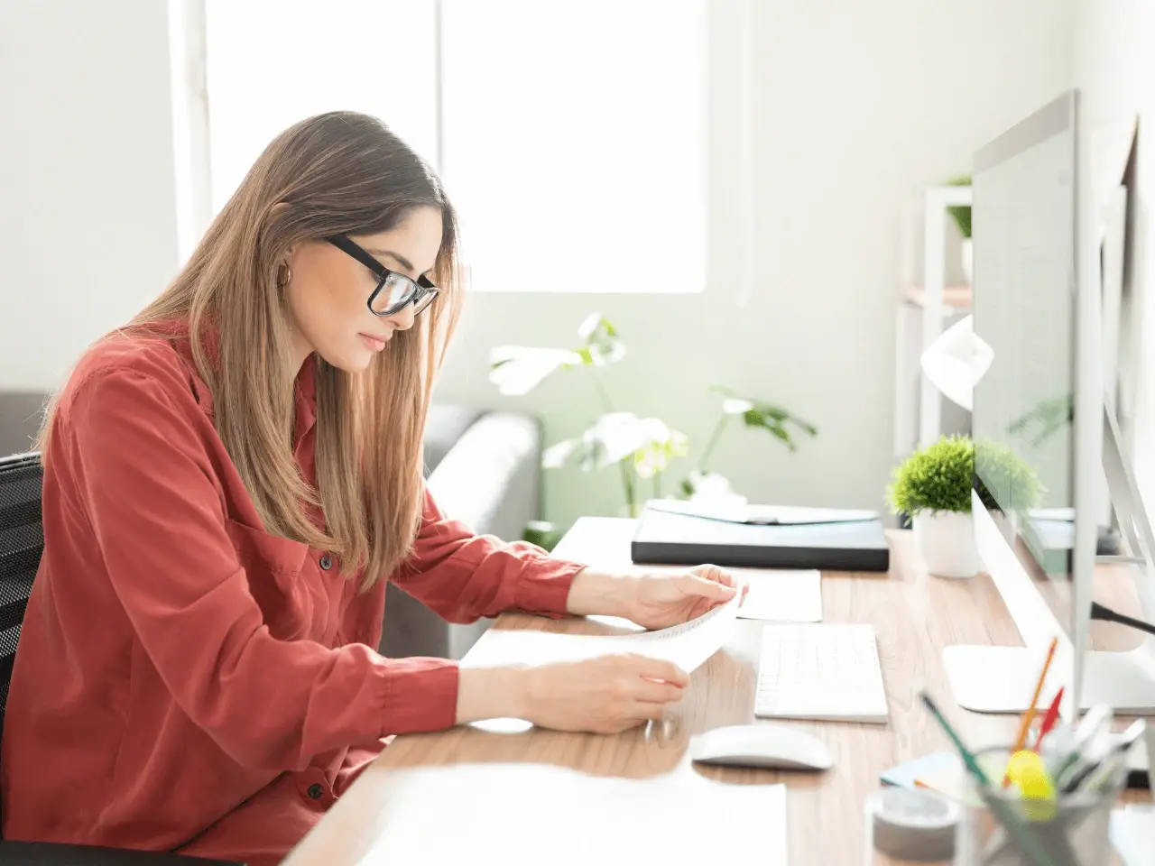 Woman in glasses working at a desk, reviewing printed documents while using a desktop computer, representing the human review process in translation quality control.