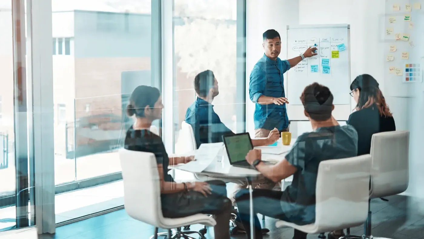 Business team in a meeting room, with one person presenting multilingual marketing strategies on a whiteboard while colleagues listen and take notes.