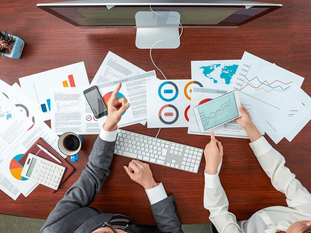 Two business professionals reviewing multilingual marketing data and global sales charts at a desk with documents, tablet, and computer.
